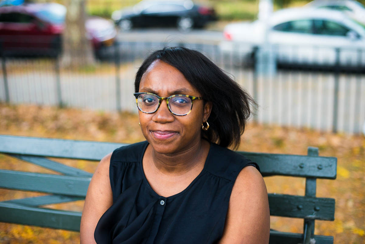 Headshot of Edwina Hay. I am seated on a park bench, wearing glasses, and a black top and the wind has blown my hair.